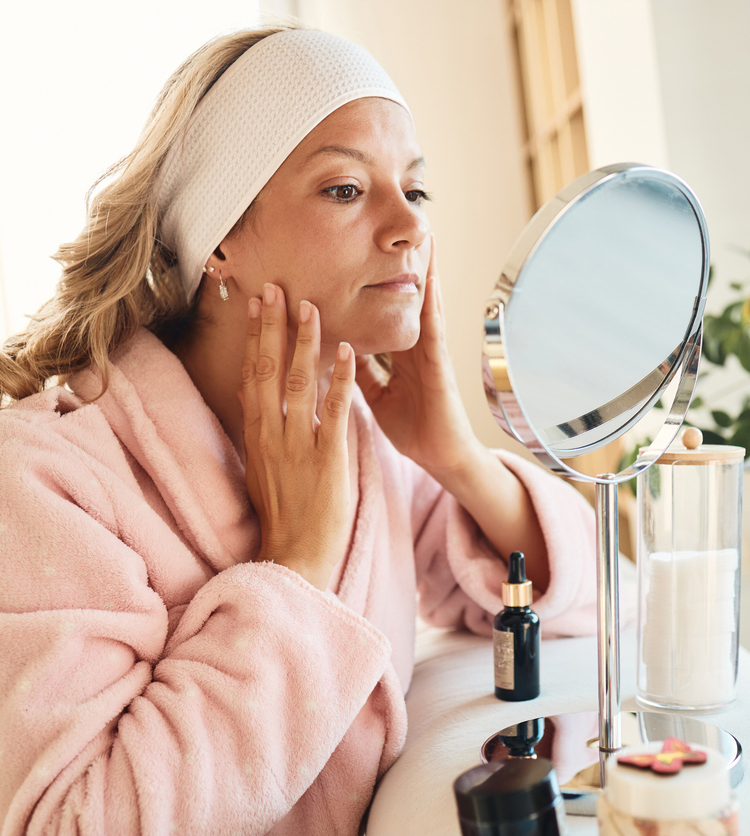 A woman applying skincare in front of a mirror wearing a pink robe.