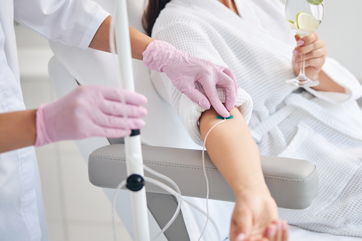 Nurse administering IV drip to a woman sitting in a robe while holding a drink.
