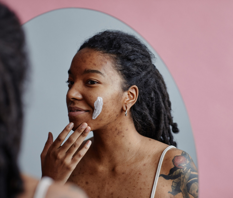 A woman applying facial cream while looking in a mirror with pink background.