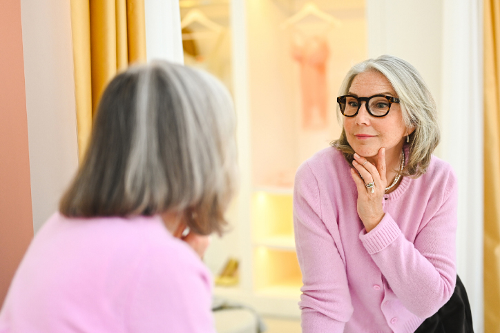 Woman in pink sweater and glasses admires reflection in a mirror indoors.