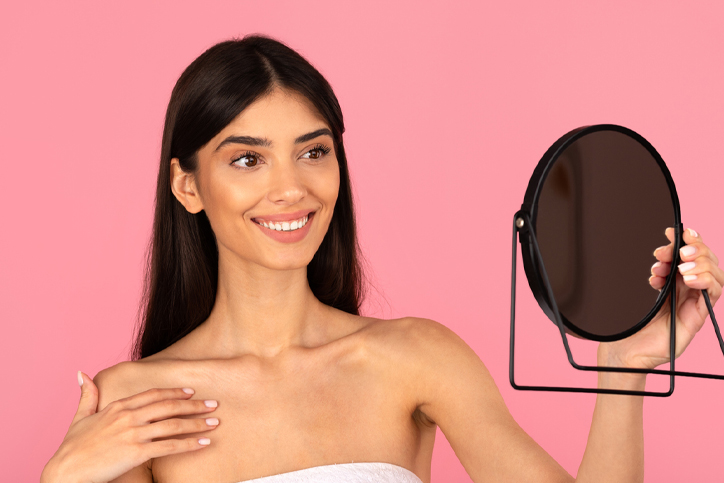 A woman smiling and holding a mirror against a pink background.