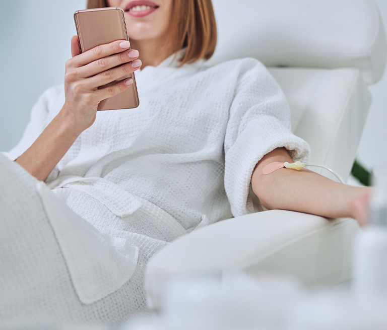 A woman in a spa robe receiving IV therapy while using a smartphone.