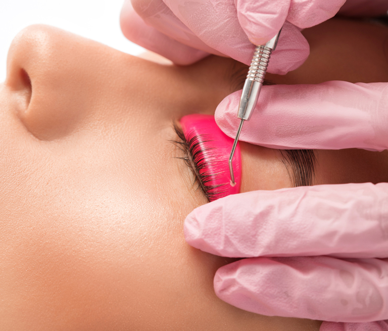 Eyelashes being curled with a pink rod and tool by gloved hands in beauty treatment.