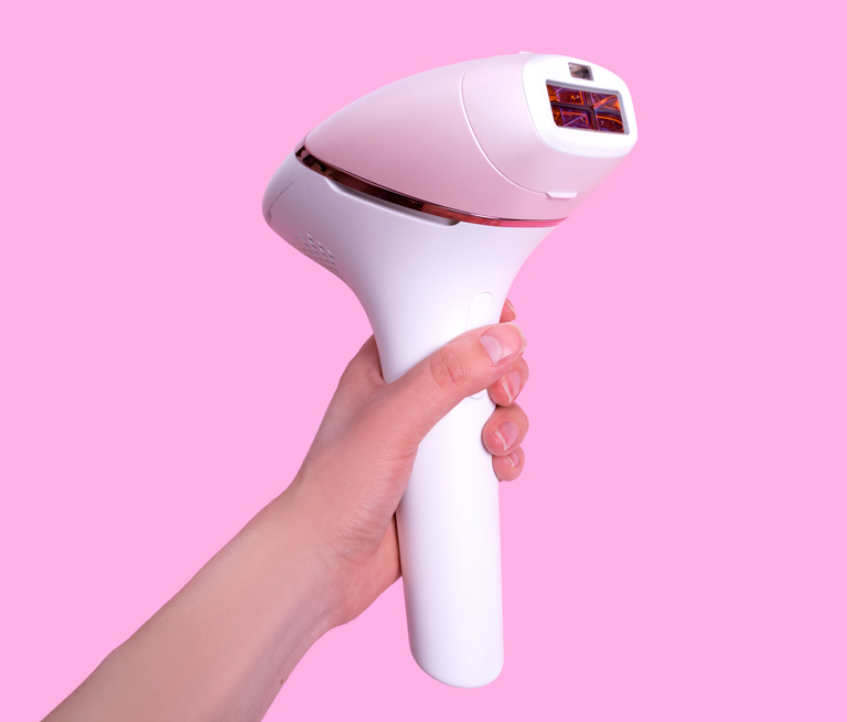 A hand holding a white IPL hair removal device against a pink background.