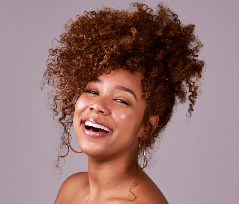 Smiling woman with curly hair and hoop earrings against a neutral background.