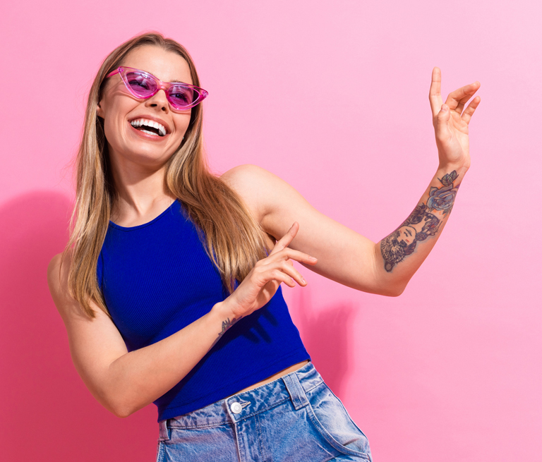 A woman in pink sunglasses, blue top, and jeans, smiling against a pink background.