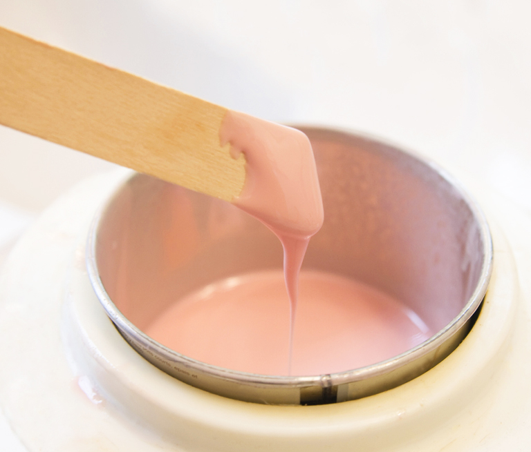 Pink wax being stirred in a warmer with a wooden spatula.