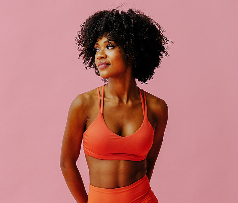 A woman in orange activewear stands against a pink background.