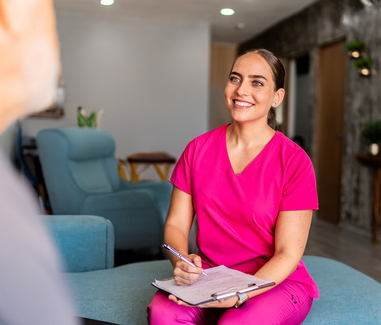 A woman in pink scrubs smiling and taking notes in a medical office.
