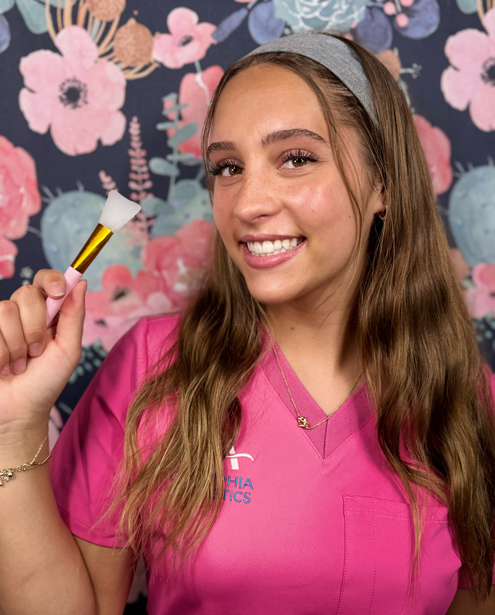 A woman in a pink uniform holding a brush, smiling in front of floral wallpaper.